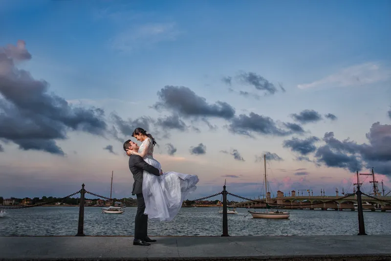 Groom lifts bride on bayfront with Bridge of Lions at sunset