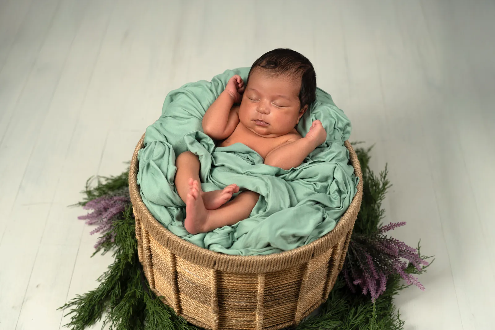 Newborn in wicker basket with sage green wrap and lavender accents