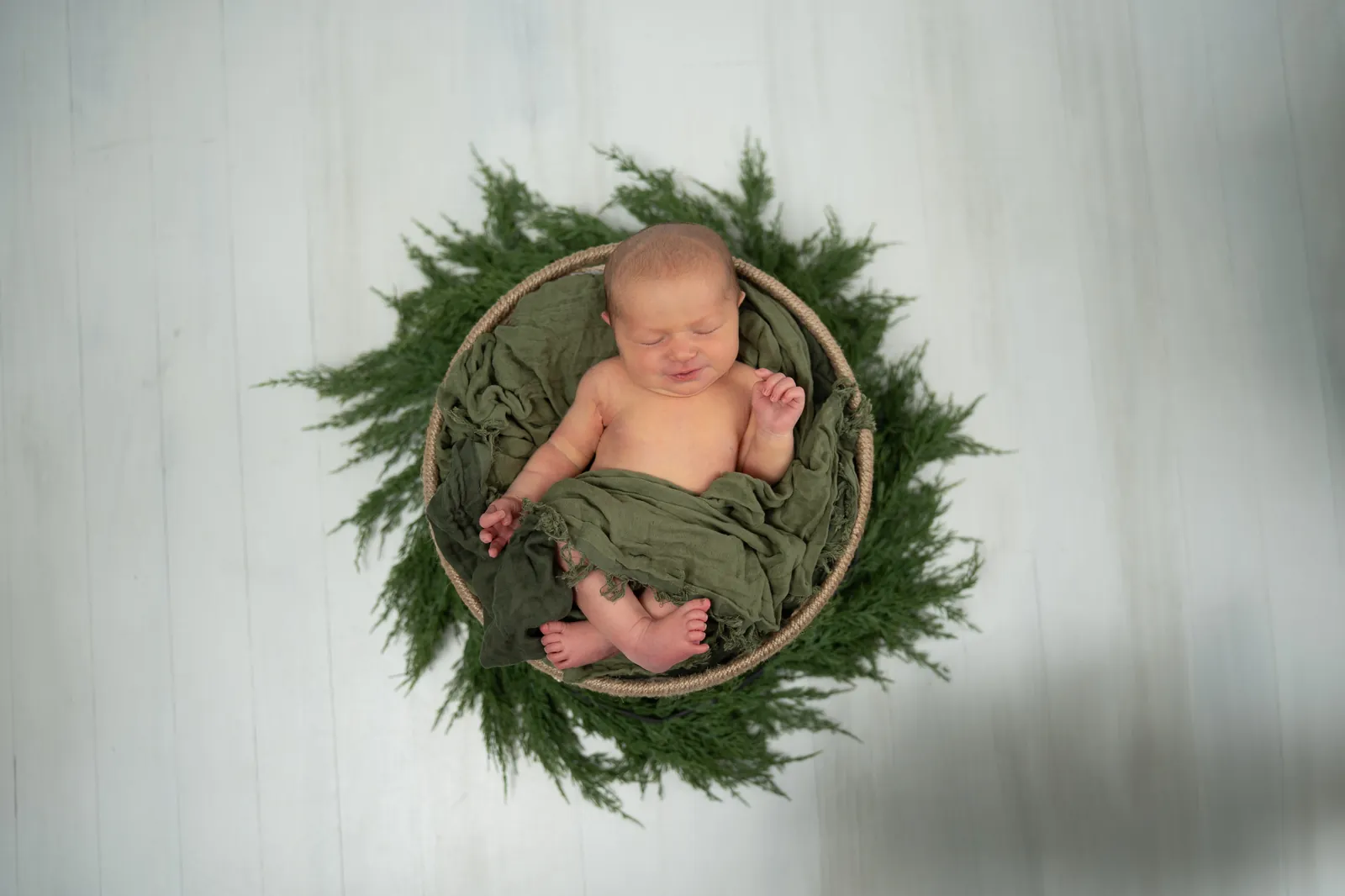 Newborn baby in wicker basket with olive wrap surrounded by greenery