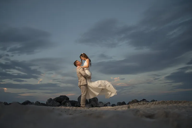 Groom lifts bride on beach at twilight