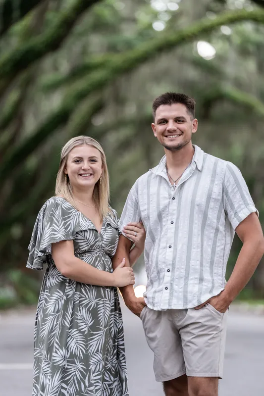 Abby and Gage stand together smiling under sprawling oak trees, Abby in a patterned dress and Gage in a striped shirt