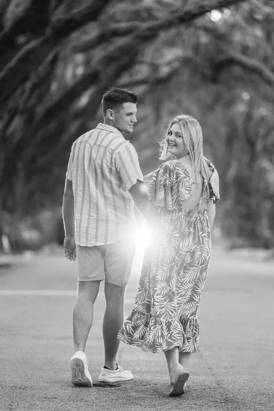Black and white image of Gage and Abby facing each other on a shaded avenue lined with massive oak trees, sunlight filtering