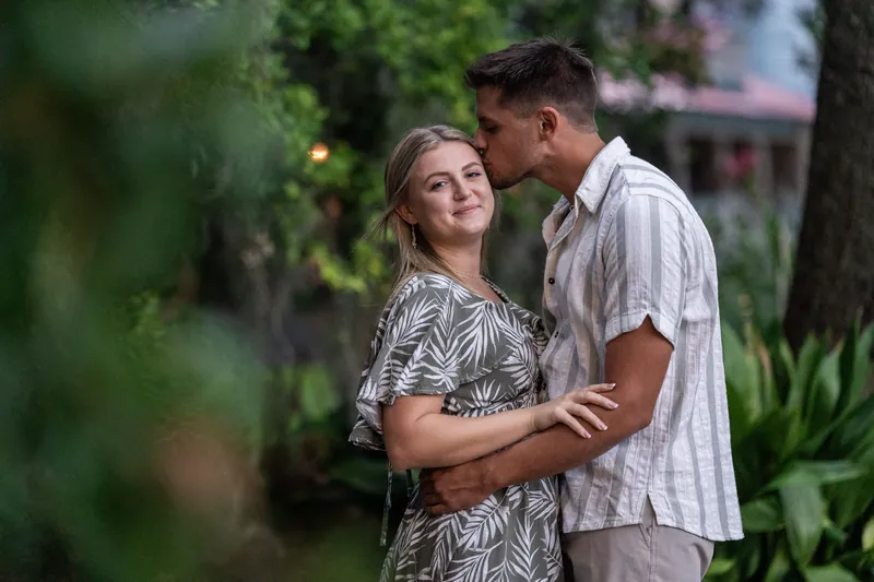 Abby looks up at Gage while standing close together in dappled green shade, both wearing patterned and striped tops.