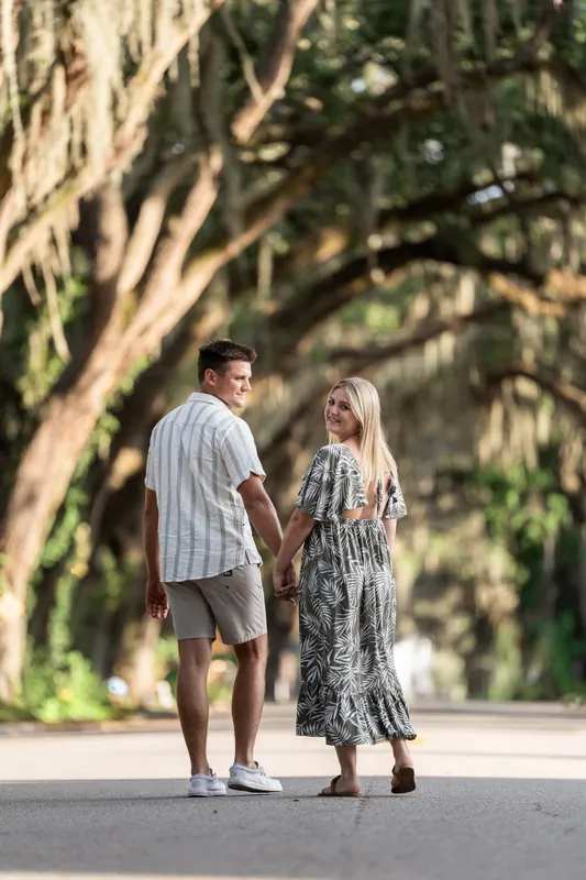 Gage and Abby stand apart on a sunlit tree-lined street, the massive oak branches creating dramatic shadows across