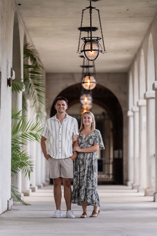 Gage and Abby walk hand in hand down a long covered corridor at The Loggias, with hanging lanterns and white columns