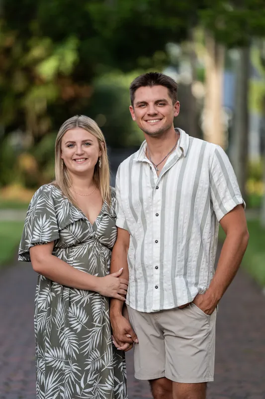 Abby in a botanical print dress and Gage in a striped shirt smile warmly at the camera on a tree-lined residential street