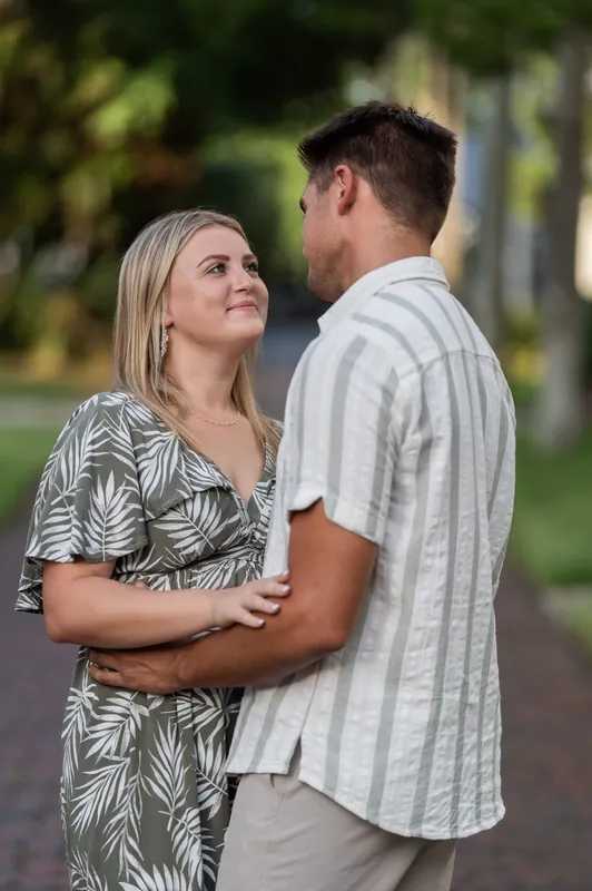 Gage gently holds Abby close as they embrace on Palm Row, with soft green foliage blurred in the background.