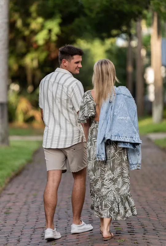 Gage and Abby walk hand-in-hand down a tree-lined brick path at Palm Row in St. Augustine, with Abby wearing a patterned