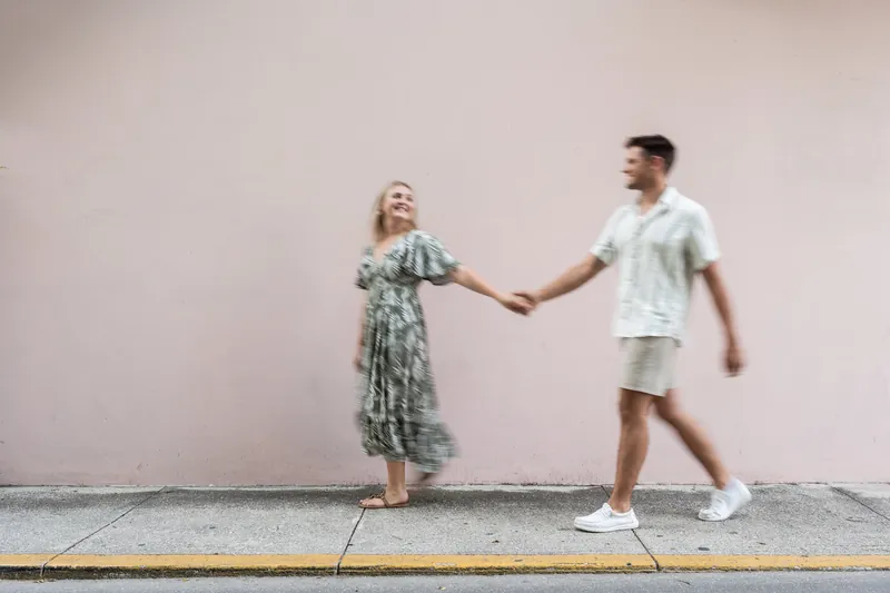 Abby and Gage hold hands while standing against a soft pink wall in downtown St. Augustine, both smiling at the camera.