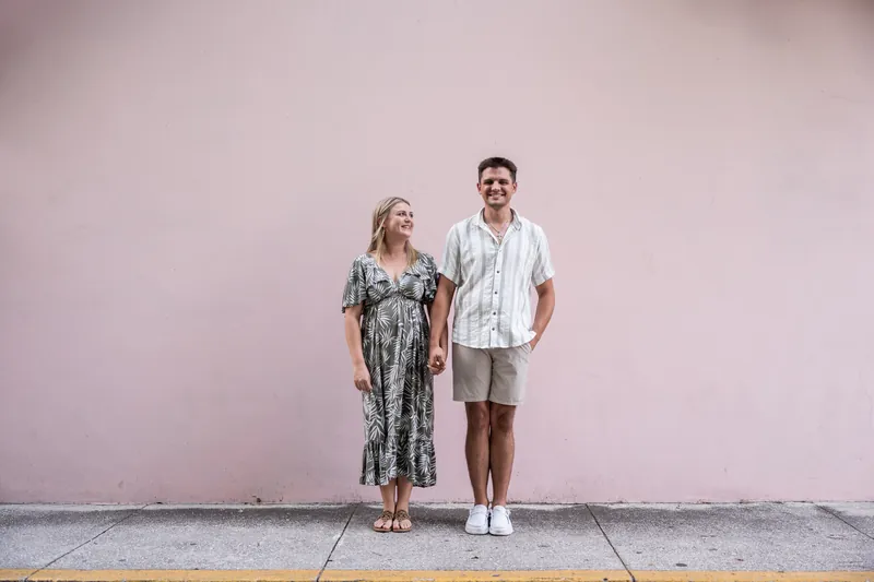 The couple stands side-by-side against a pale pink wall, with Abby in a patterned dress and Gage in a striped shirt