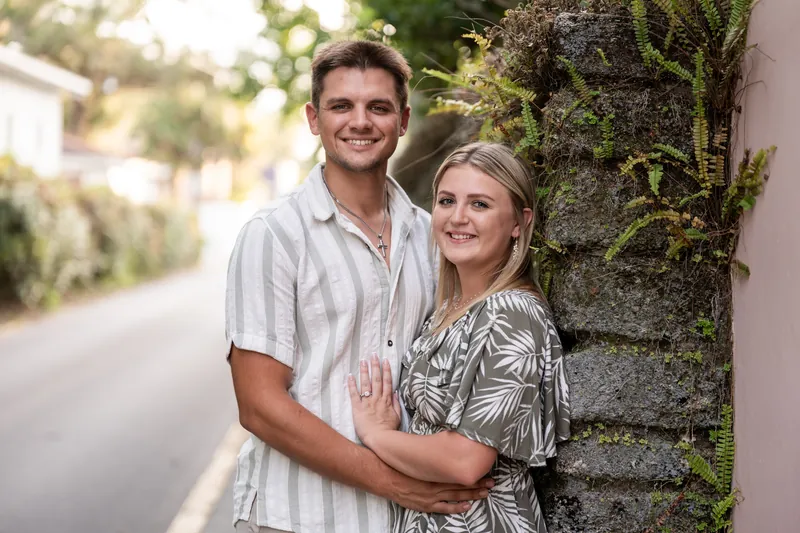 Gage smiles at the camera with his arm around Abby, who wears a palm-print top against a moss-covered stone wall.