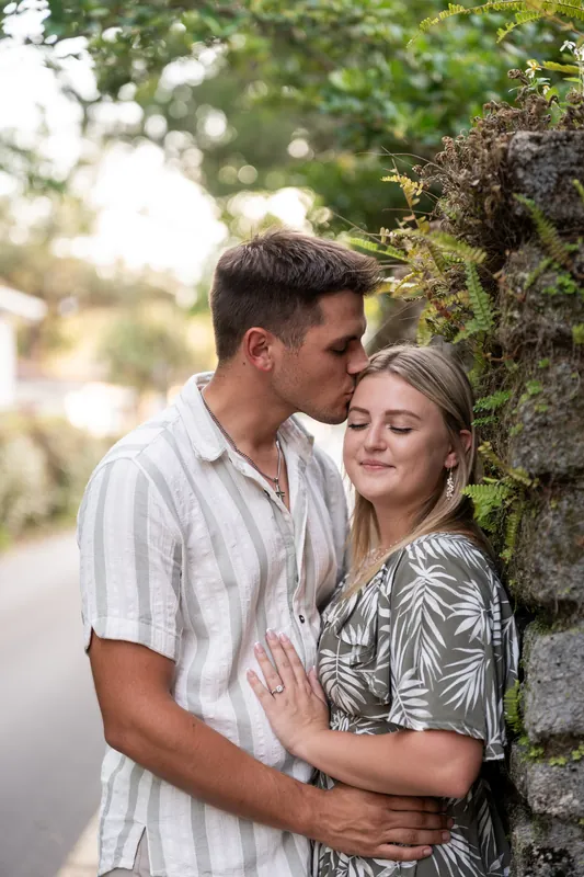 Gage leans in to kiss Abby's temple as she gazes ahead, both framed by ivy-covered brick and soft natural light.