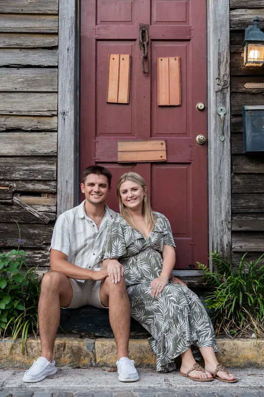 Abby and Gage sit on a step in front of a weathered red door framed by wooden siding in Downtown St Augustine.