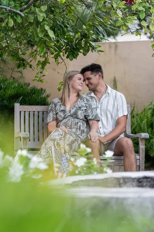 The couple sits together on a white bench beneath leafy vines, stealing a kiss in a quiet courtyard garden.