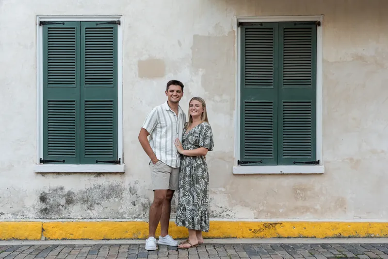 Gage and Abby stand close against a whitewashed building with green shutters and a yellow base stripe in Downtown St