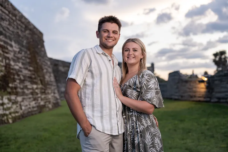 Gage wraps his arm around Abby as they smile at the camera with the stone fortification and golden sunset light behind them.