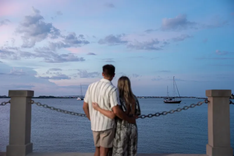 Gage and Abby embrace at the St. Augustine Bayfront railing, watching sailboats on the water at twilight.
