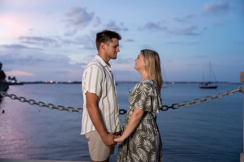 Gage holds Abby close as they face each other by the bayfront chain railing, the water and moored boats glowing in pink
