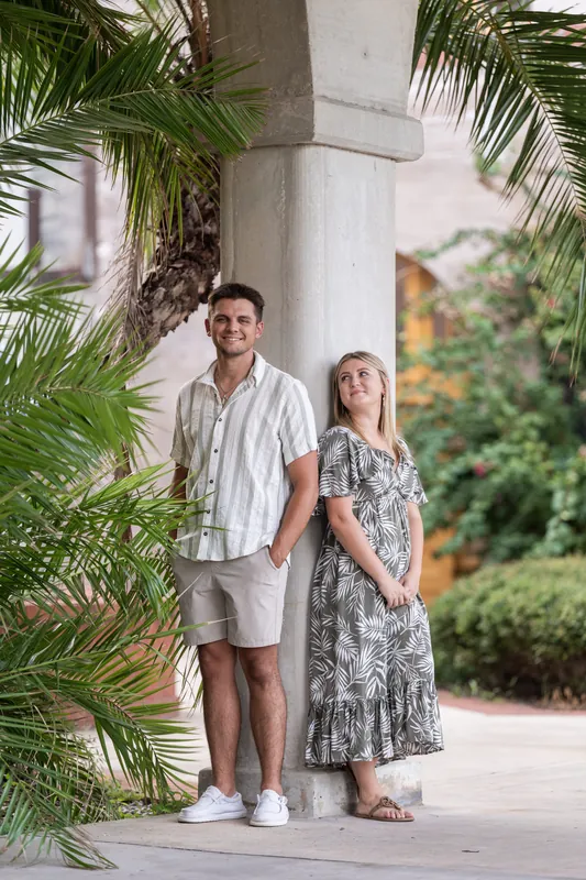 Gage and Abby pose together under a white column at The Loggias, surrounded by lush palm fronds in a tropical colonnade