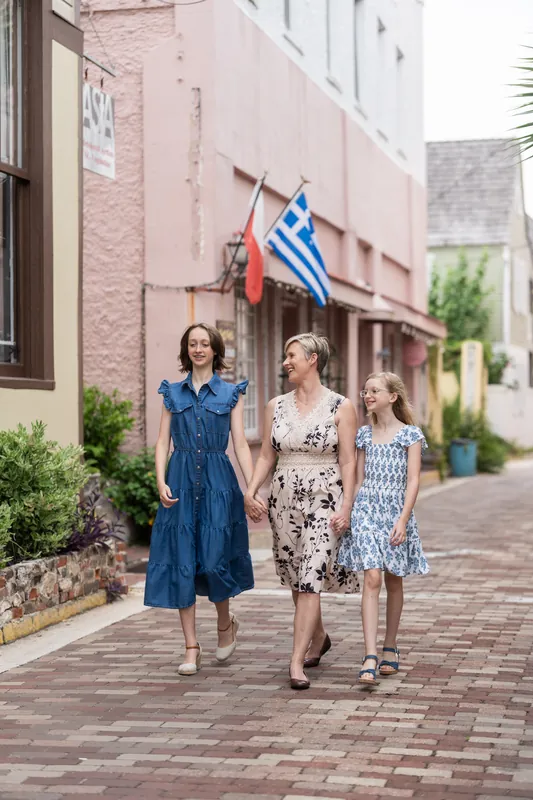 Three people walk down a brick street past a pink building with a Greek flag hanging on its facade in Downtown St Augustine.