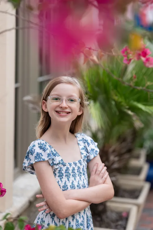 A young person in a blue patterned dress smiles at the camera with pink flowers and historic buildings blurred behind them.