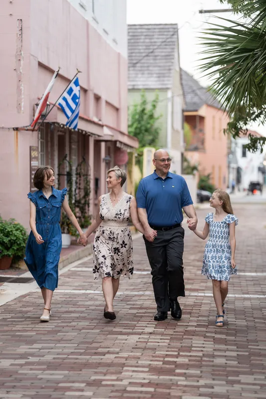 Four people stroll down a brick street past colonial buildings with a Greek flag visible on a pink facade in Downtown St Augustine.