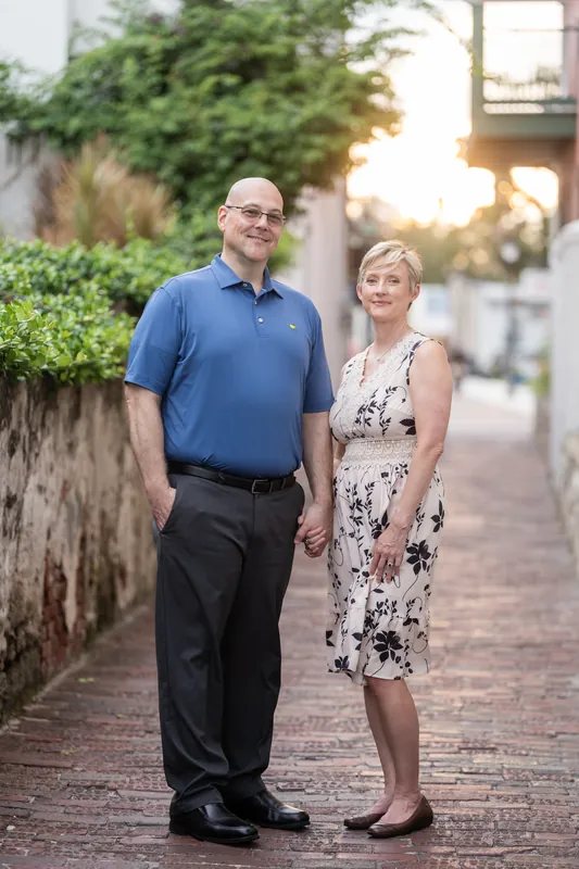 Abigail & Frank hold hands while strolling a quiet brick alley framed by stone walls and flowering vines at sunset.