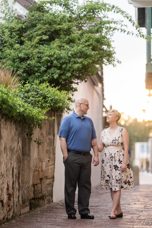 The two walk side-by-side along a tree-lined brick pathway with historic architecture and green foliage in Downtown St. Augustine.