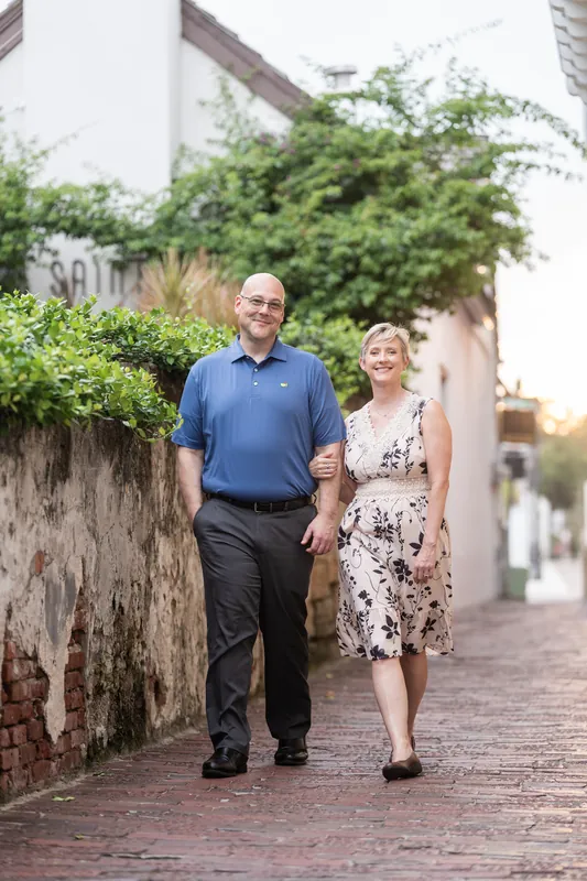 Abigail & Frank stroll together down a brick street bordered by lush vegetation and aged stone walls at dusk.