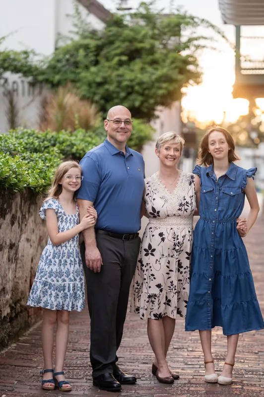 A man in a blue shirt stands with three females—ranging from child to adult—on a brick pathway lined with ivy and historic buildings.