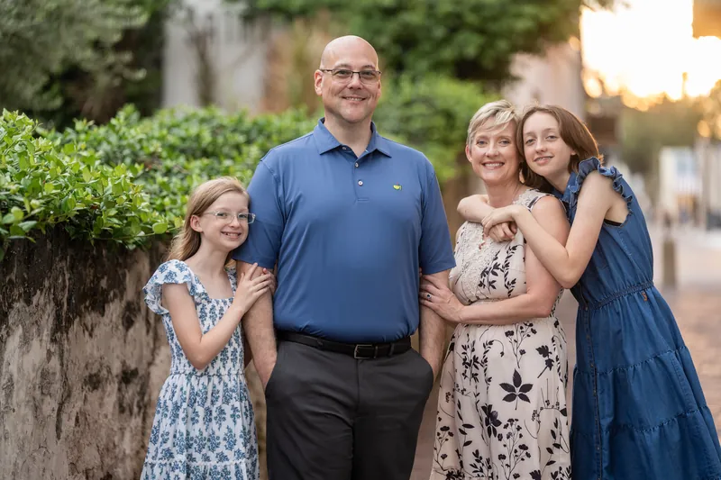 A man in a blue polo and three females in varied print dresses stand together on a brick-lined Downtown St Augustine street at golden hour.