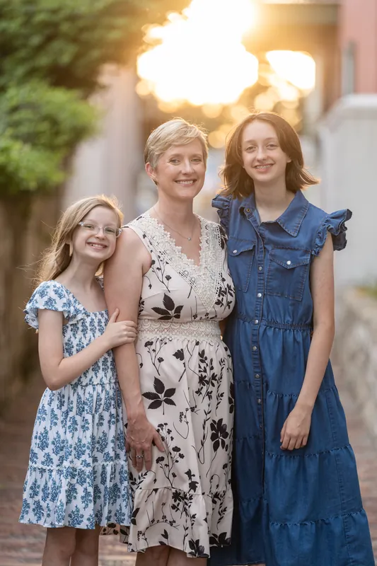 Three females in blue and floral dresses smile closely together against a softly lit Downtown St Augustine building at sunset.