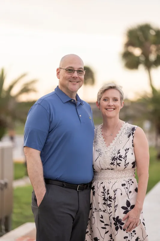 Two adults pose side-by-side on a palm-lined Downtown St Augustine pathway with warm evening light behind them.
