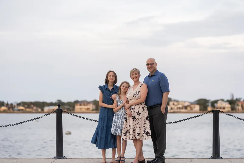 Four people stand together by a waterfront railing in Downtown St Augustine, two in blue, one in a patterned dress, under a cloudy sky.