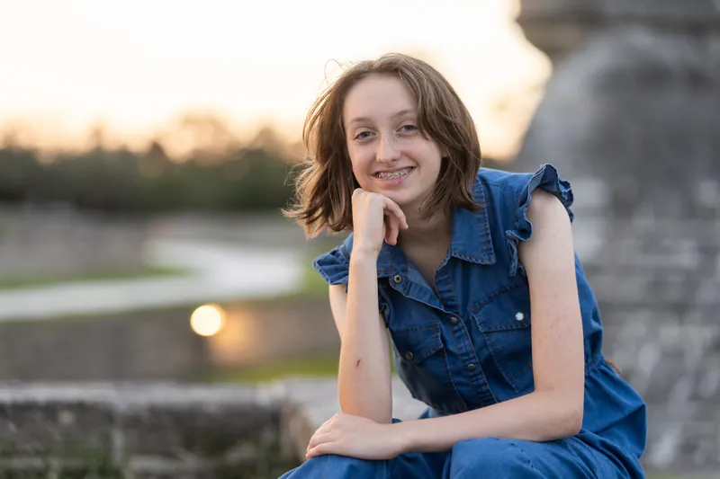 A young woman in a denim shirt smiles at the camera, posed near water at sunset in Downtown St Augustine.
