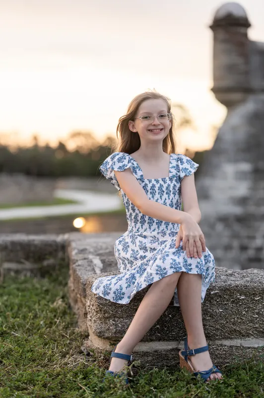 A girl in a blue-and-white floral dress sits on a stone ledge, gazing off-camera with golden hour light behind her.