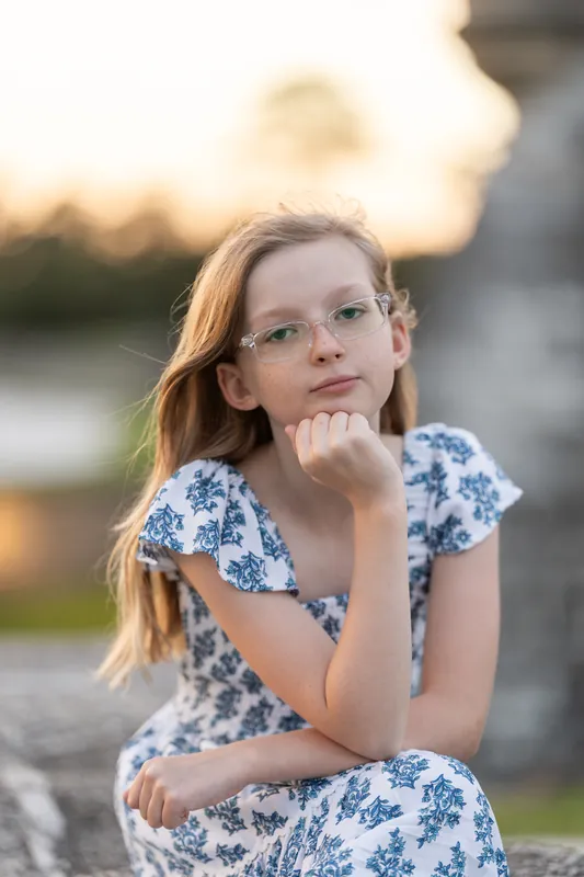 A girl wearing glasses and a patterned dress rests her chin on her hand in a thoughtful pose near a historic structure.