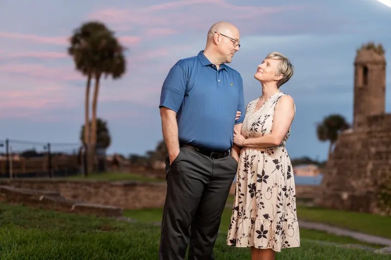 Abigail and Frank stand together at dusk in Downtown St Augustine, with palmetto trees and historic ruins framing the twilight sky behind them.
