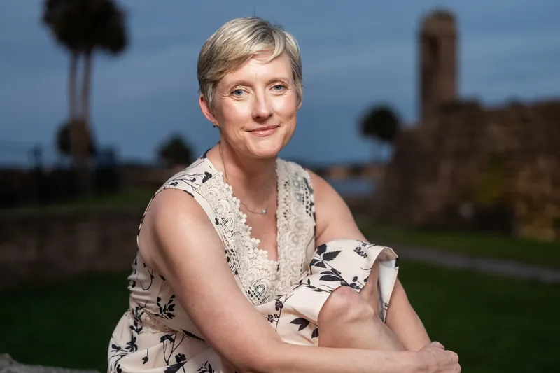 A person in a floral dress smiles toward the camera with historic St Augustine landmarks and palmettos visible across the evening landscape.