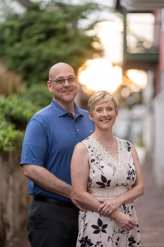 A man in a blue polo shirt and a woman in a floral dress walk arm-in-arm down a brick street with historic pink buildings behind them.