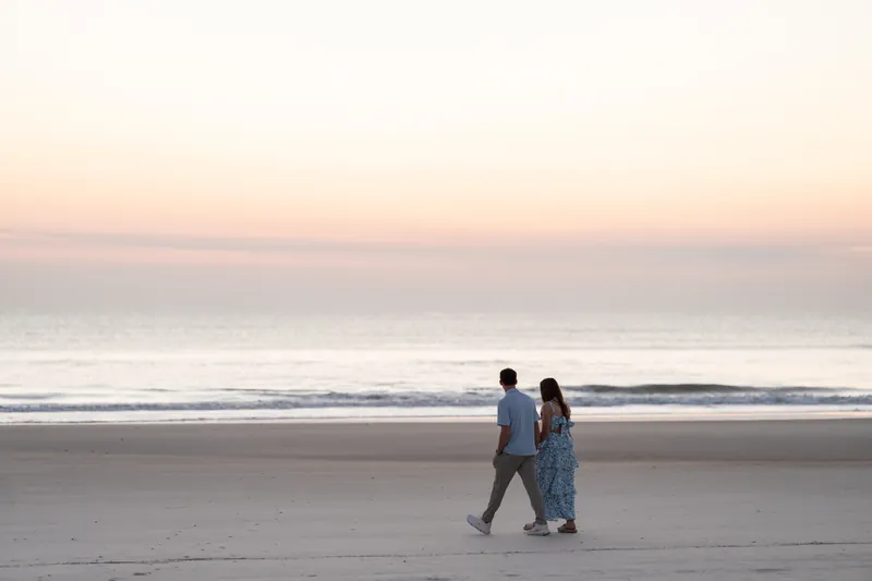 Alex and Teigan walk hand in hand along a wet sandy beach at sunset with calm ocean waves in the background.