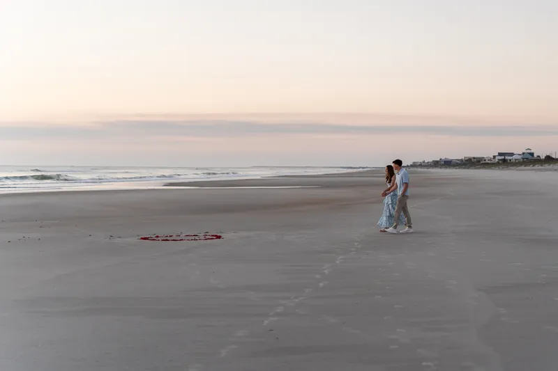 Teigan stands facing Alex as he proposes on the beach during golden hour with the ocean and distant shoreline in view.