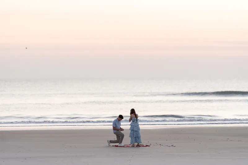 Alex kneels before Teigan on the beach with a ring visible, proposing at sunset with soft waves rolling in behind them.