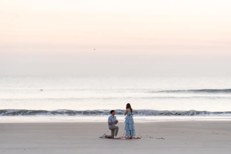 Teigan embraces Alex after accepting his proposal on the beach at sunset, with the ocean and pastel sky behind them.