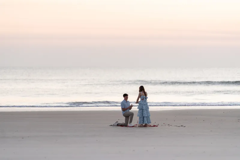 Alex and Teigan embrace on the beach at sunset, standing on red rose petals with calm ocean waves behind them.