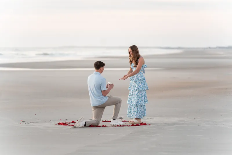 Alex proposes to Teigan on one knee in his light blue shirt as she stands in her patterned blue dress, with rose petals marking the proposal spot on the sand.