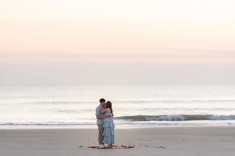 Teigan and Alex stand together on the beach at dusk, embracing on a bed of red rose petals with the ocean behind them.