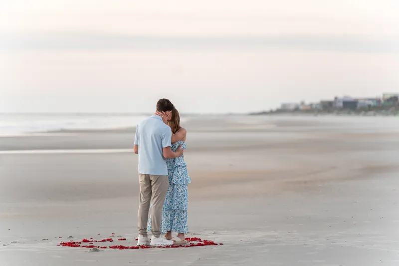 Alex wraps his arm around Teigan from behind as they stand together on the beach with rose petals at their feet and the shoreline visible in the background.