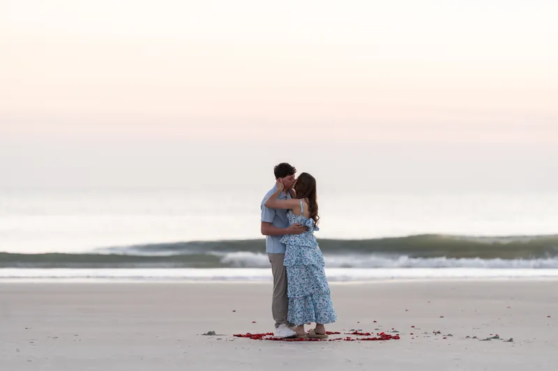 Alex and Teigan embrace tightly on the beach while standing on scattered red rose petals, with waves visible behind them in the twilight.