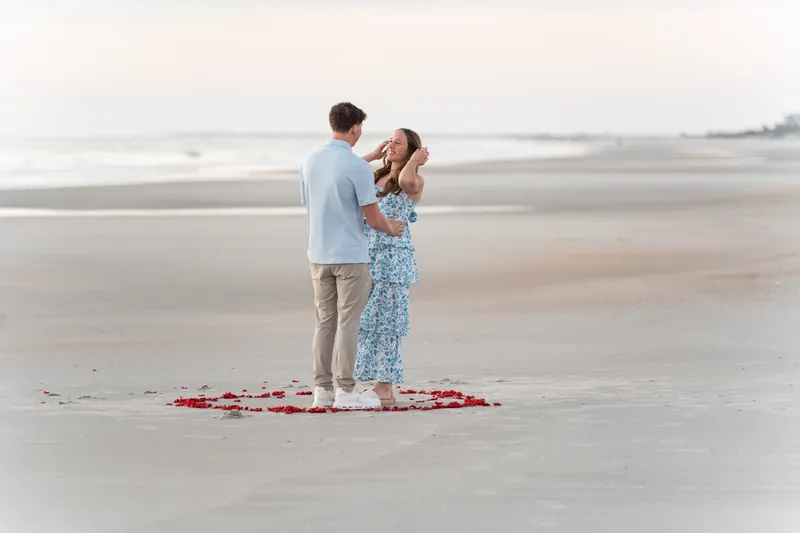 Alex proposes to Teigan on a wide beach at sunset, standing together in the center of a heart made from rose petals.